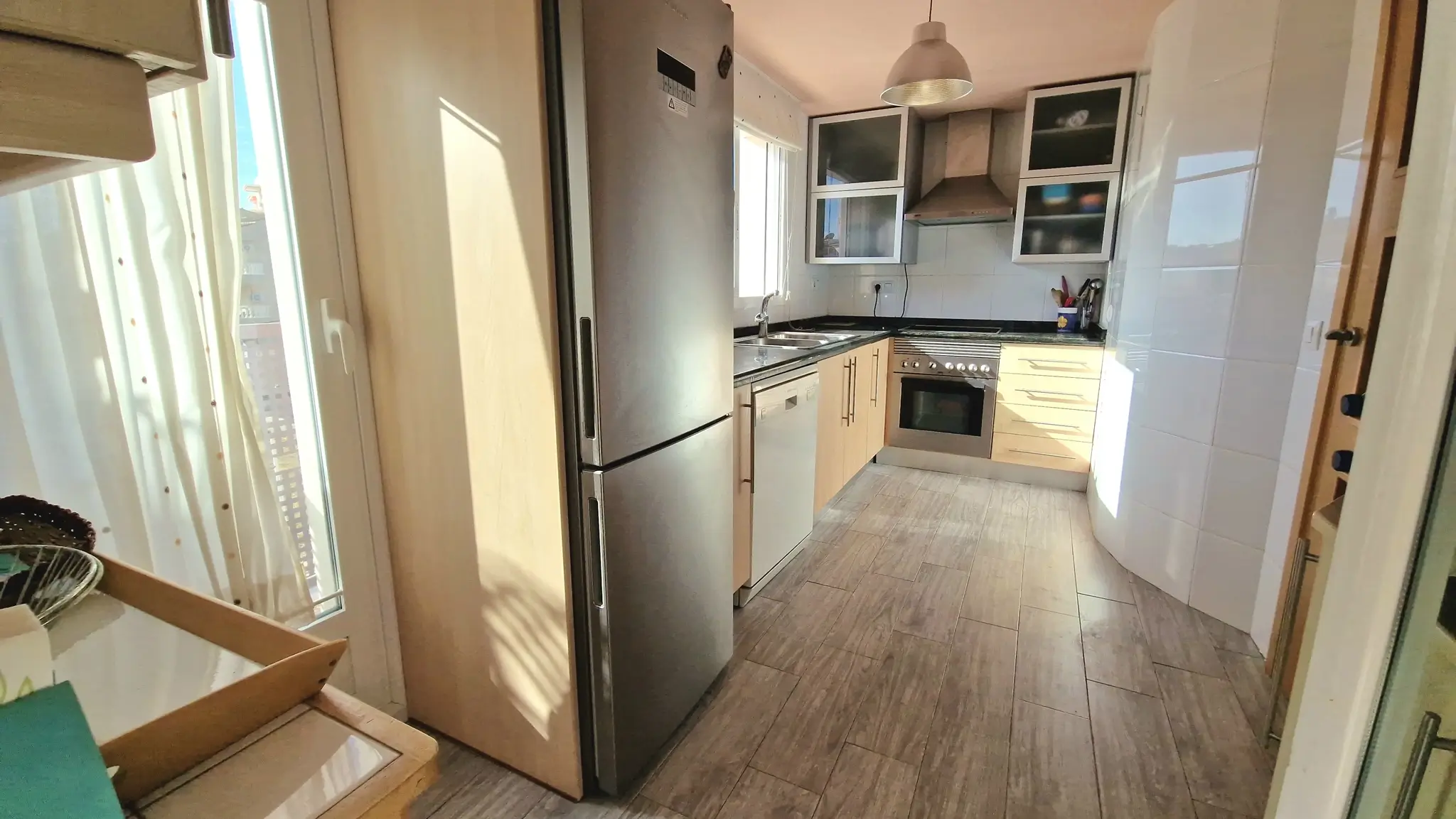 Moderne keuken met roestvrijstalen apparatuur, lichte houten kasten en een keramische tegelvloer met houtlook, verlicht door natuurlijk daglicht.