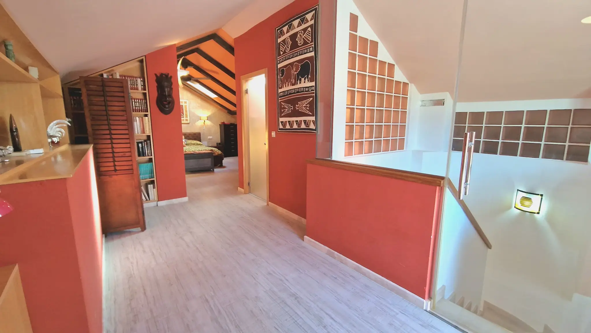 Overloop op de bovenverdieping met terracotta wanden en een glazen balustrade. Doorkijk naar een slaapkamer met schuin plafond, donkere houten balken en ingebouwde boekenkasten.