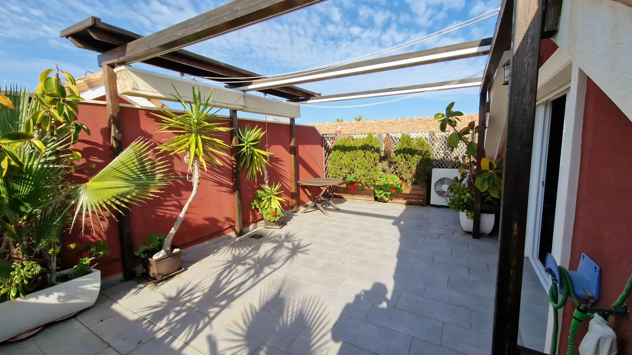 Privéterras met grijze tegels, een donkerhouten pergola en uitschuifbare zonneschermen. Voorzien van terracotta muren, diverse palmen in potten en een houten tafel.