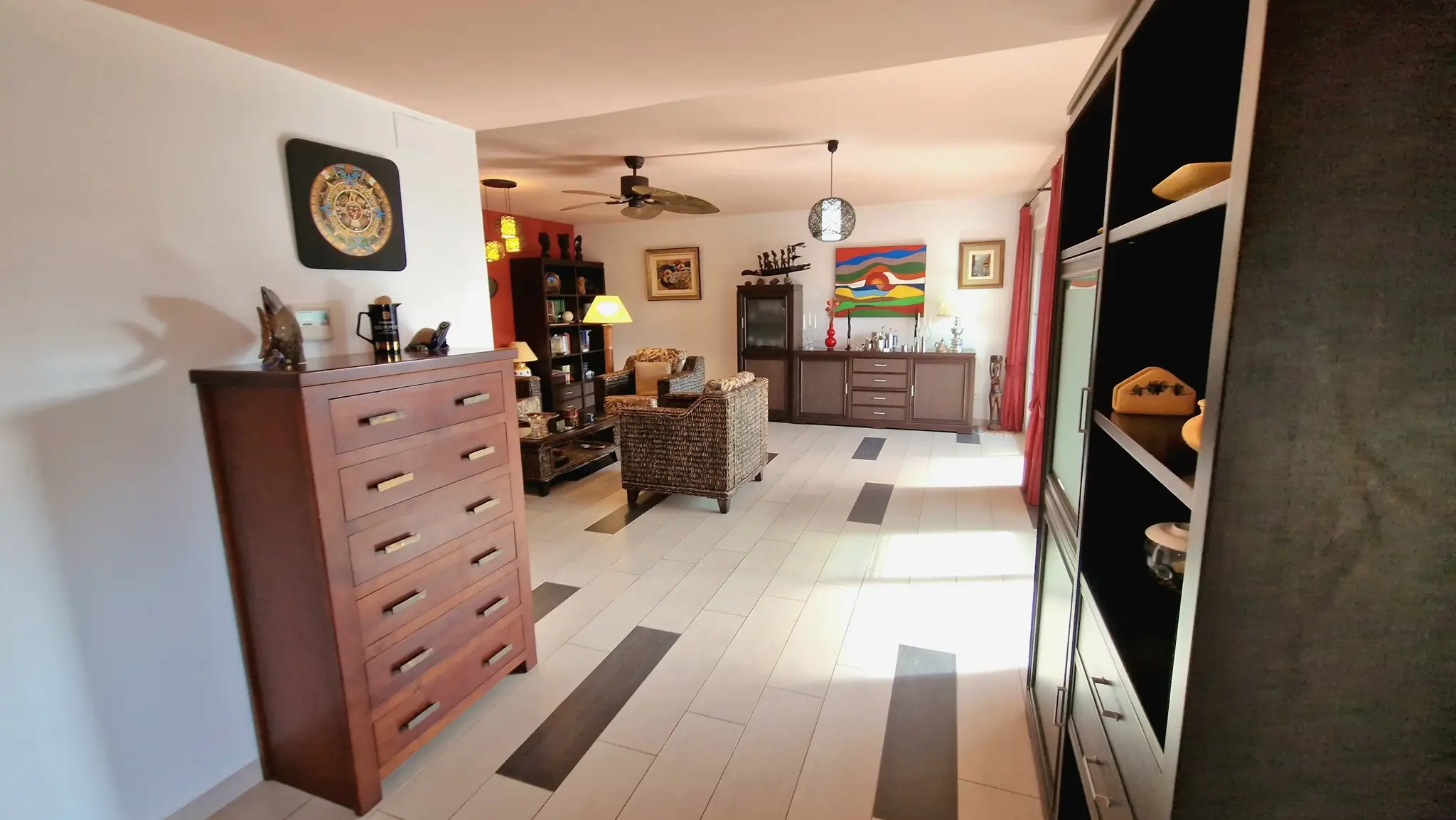Ruime woonkamer met lichte tegelvloer voorzien van donkere accenten, rieten fauteuils, een houten ladekast en een centrale plafondventilator.