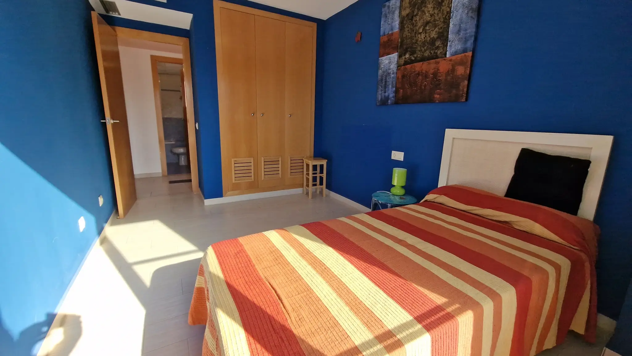 Slaapkamer met diepblauwe muren, een houten inbouwkast en tegelvloer. De ruimte is voorzien van een eenpersoonsbed en biedt directe toegang tot een aangrenzende badkamer.