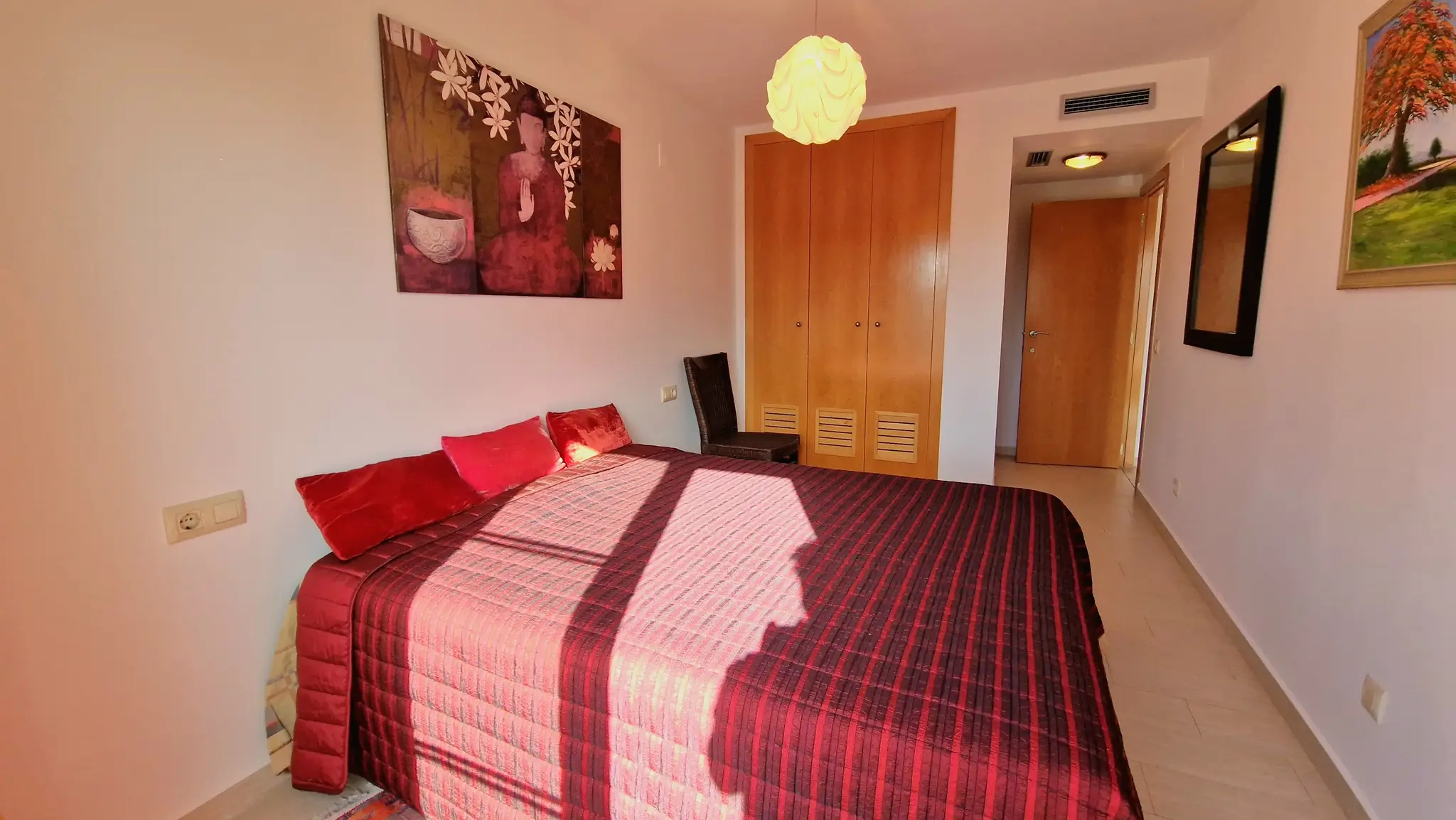 Slaapkamer met tweepersoonsbed, bordeauxrood gestreept beddengoed, houten inbouwkasten en oosterse wanddecoratie met een Boeddha-afbeelding.