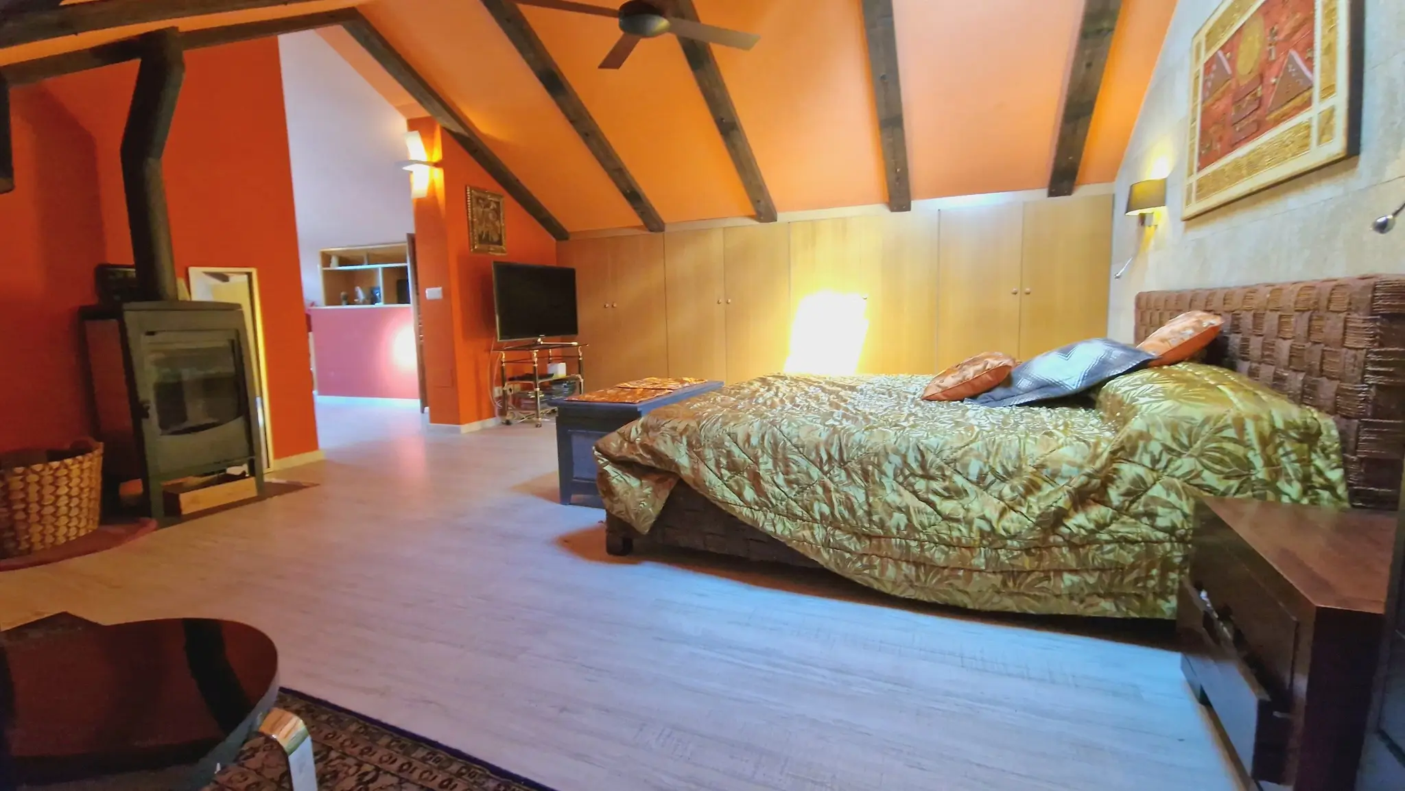 Slaapkamer onder een schuin dak met houten balken, een houtkachel en terracotta muren. Voorzien van inbouwkasten, een plafondventilator en een gevlochten hoofdbord.