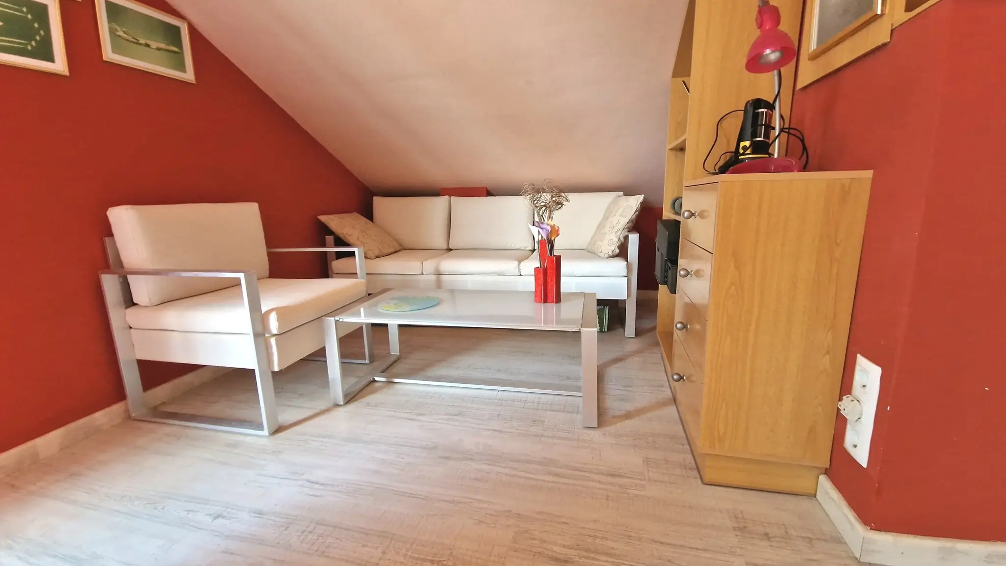 Zolderkamer met rode muren, witte zitmeubels met metalen frame, een glazen salontafel en een lichte laminaatvloer.