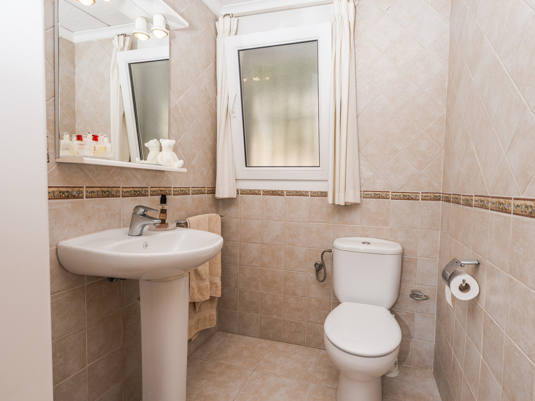 Badkamer met een kolomwastafel, wit keramisch toilet en beige betegelde wanden met een decoratieve rand en een raam voor daglicht.
