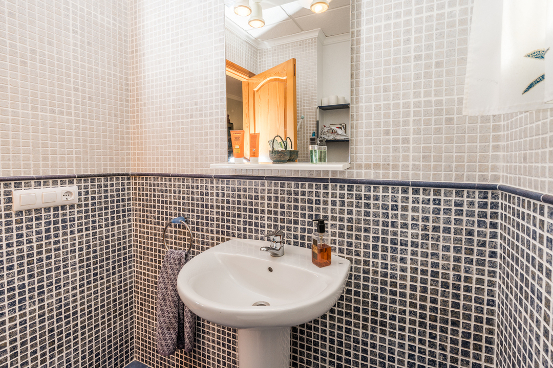 Badkamer met een witte kolomwastafel, wanden van wit-blauwe mozaïektegels en een spiegel met geïntegreerde verlichting en een planchet.