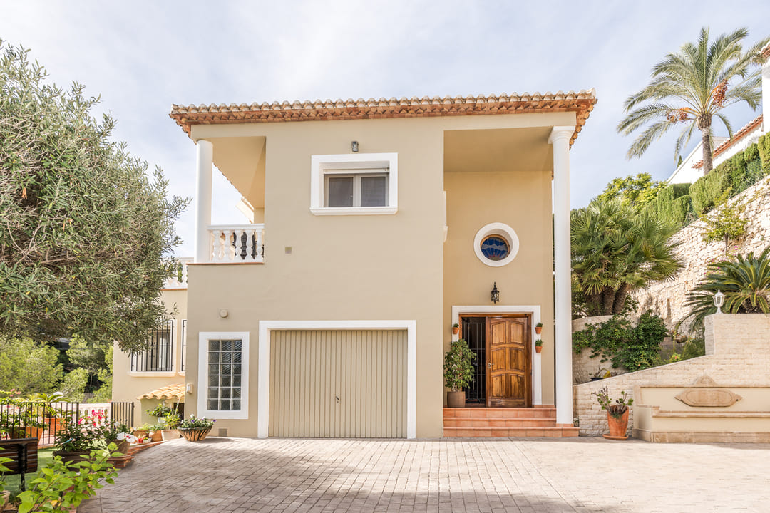 Buitengevel van een villa met twee verdiepingen, beige afwerking, geïntegreerde garage, houten voordeur en een geplaveide oprit met mediterrane beplanting.
