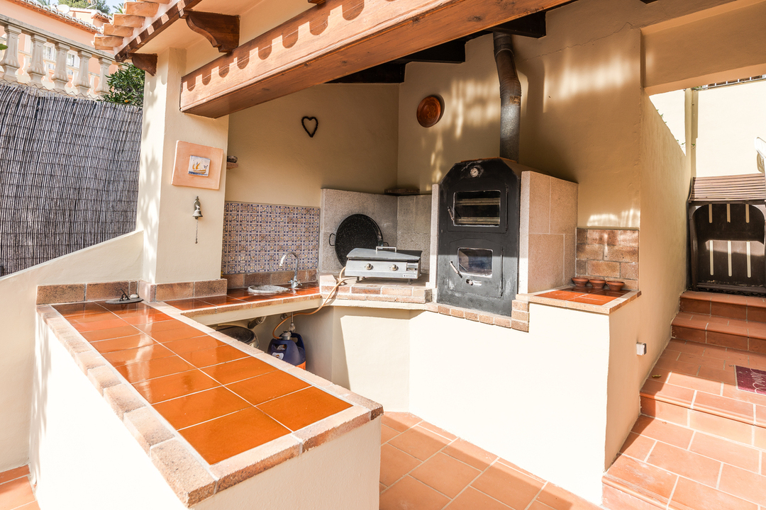 Buitenkeuken met houtoven, aanrechtbladen van terracotta tegels en een decoratieve achterwand onder een houten overkapping.