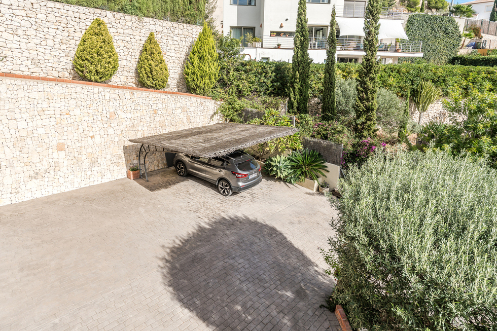 Buitenparkeerplaats met een geplaveide oprit en carport met heidedak, omgeven door een natuurstenen muur en mediterrane beplanting.