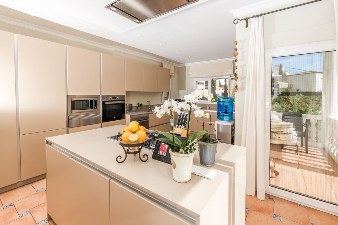 Moderne keuken met een beige kookeiland, gaskookplaat, inbouwapparatuur en terracotta vloertegels. Een glazen deur biedt directe toegang tot het terras.