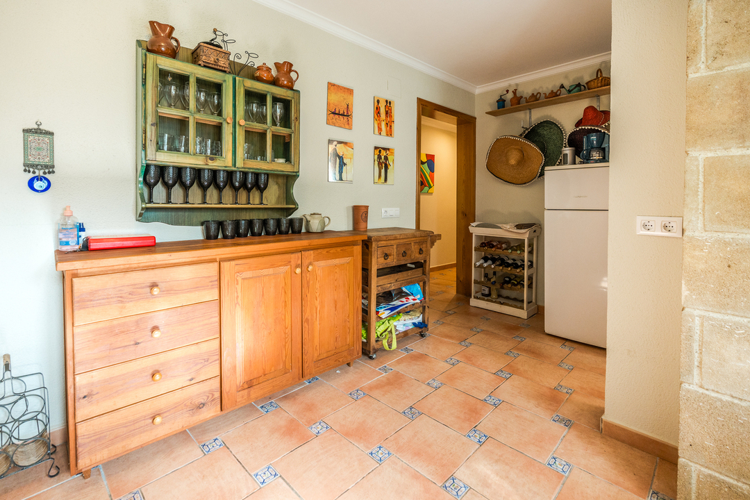 Rustiek houten dressoir en groene wandkast met glaswerk boven een terracotta tegelvloer met decoratieve details. De ruimte is voorzien van een wijnrek en een witte koelkast.