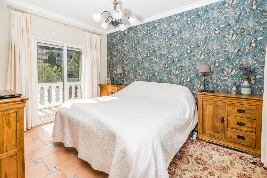 Slaapkamer met botanisch behang, houten meubels en een terracotta tegelvloer. Een schuifpui biedt toegang tot het balkon met witte balustrades.