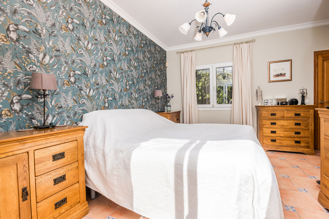 Slaapkamer met een tropische accentmuur, massief houten ladekasten en een terracotta tegelvloer met decoratieve details. Natuurlijk licht valt binnen via het raam.