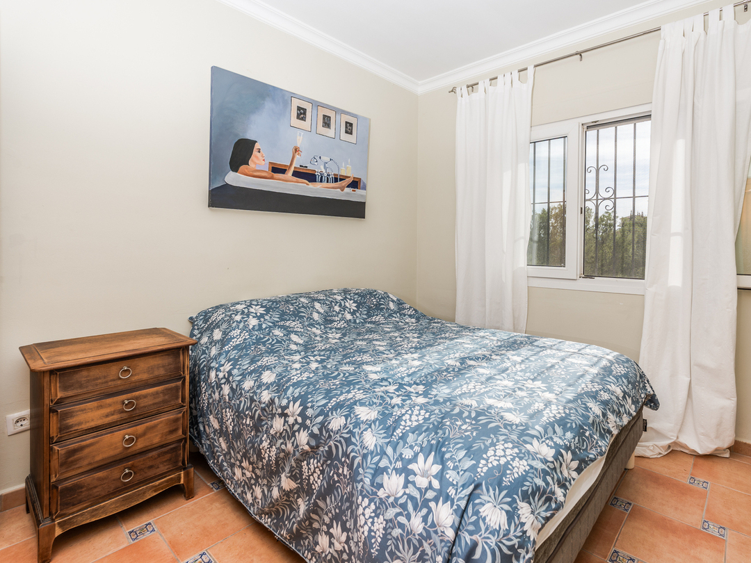 Slaapkamer met tweepersoonsbed, blauw bloemendekbed, klassiek houten nachtkastje en terracotta vloertegels met decoratieve accenten.