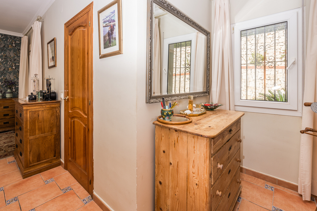Slaapkamerhoek met grenen ladekast, terracotta tegelvloer met decoratieve inzetstukken en een grote zilverkleurige spiegel naast het raam met tralies.
