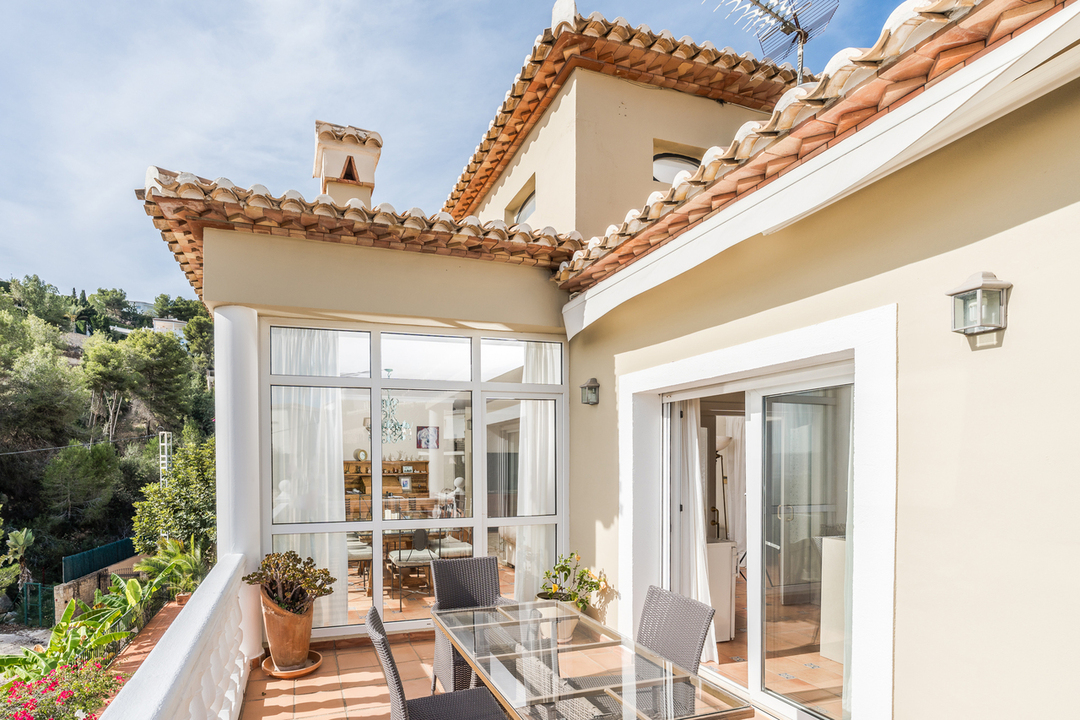 Terras met terracotta tegels, een glazen eettafel en witte balustrade. Grote raampartijen tonen een serre, omgeven door mediterrane architectuur en groene heuvels.