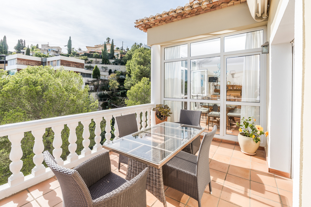 Terras met terracotta tegels, witte balustrade en glazen eettafel. Het biedt uitzicht op de groene heuvelrug en de omliggende mediterrane bebouwing.