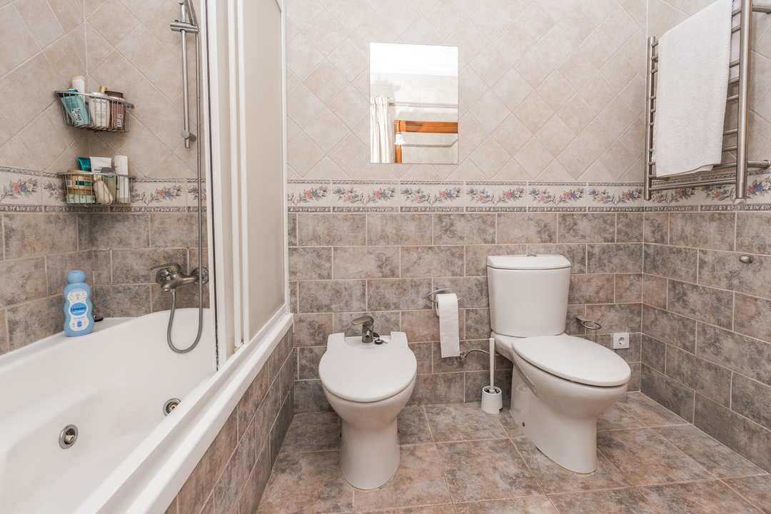 Volledige badkamer met ligbad, bidet en toilet. De wanden zijn bekleed met aardetinten tegels, een decoratieve bloemenrand en een chromen handdoekradiator.