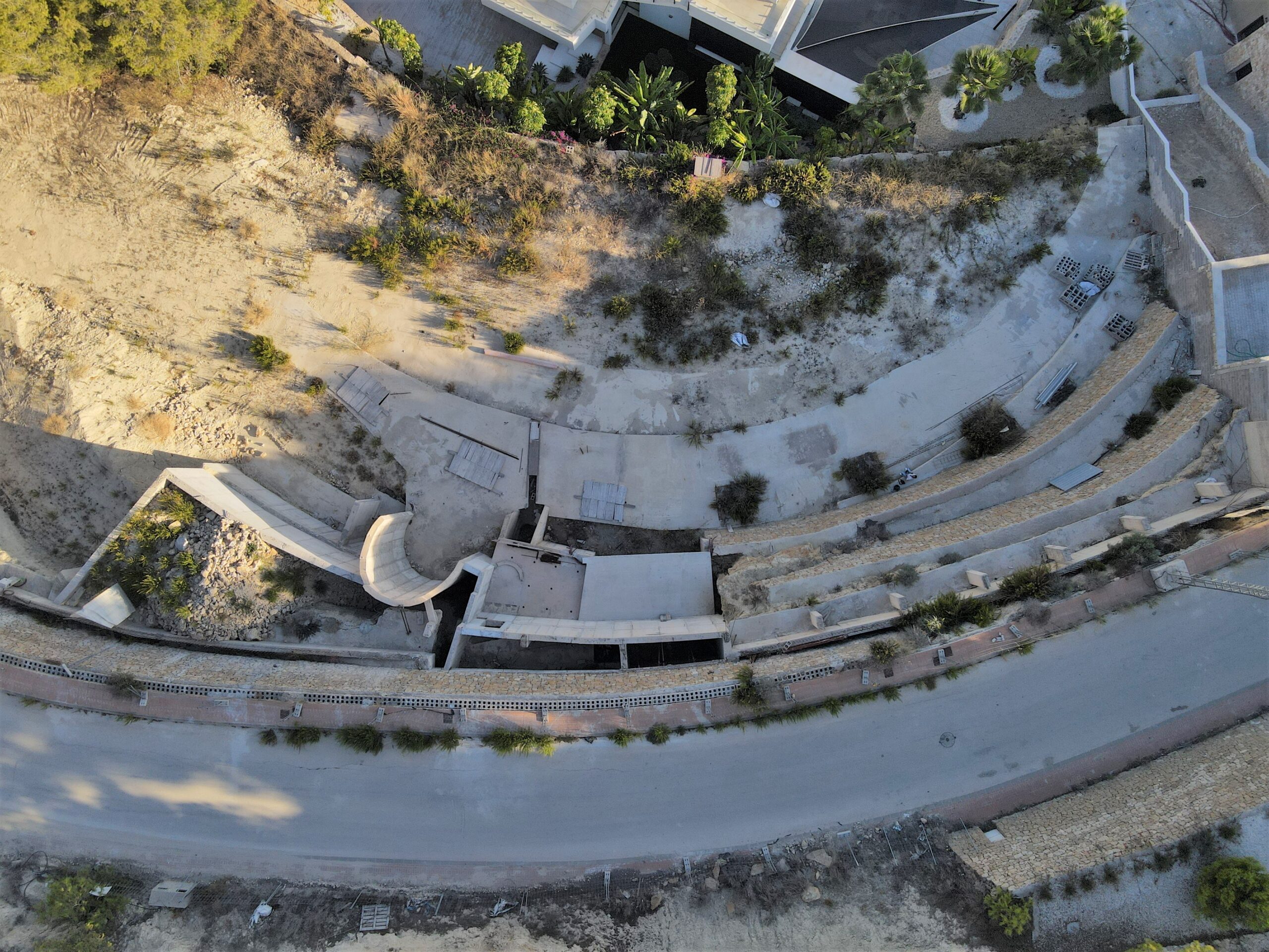 Luchtfoto van een bouwterrein met terrasvormige betonnen funderingen en stenen keurmuren, geïntegreerd in het glooiende landschap.