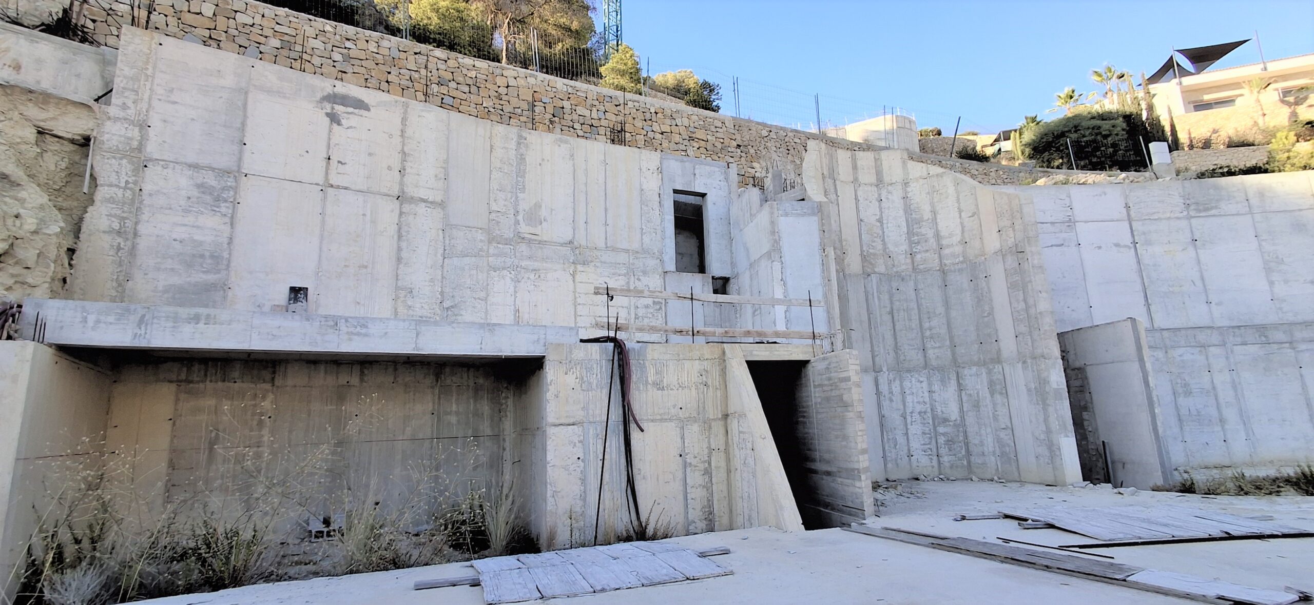 Structureel aanzicht van de gewapend betonnen steunmuren, met geïntegreerd natuursteen op het bovenste niveau en architectonische openingen op een hellend terrein.