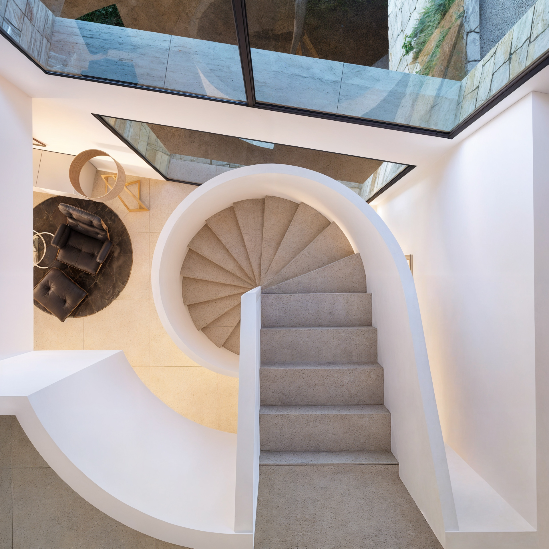 Spiraalvormige stenen trap met witte gebogen balustrades van bovenaf gezien, inclusief grote raampartijen en een zithoek met een leren fauteuil.