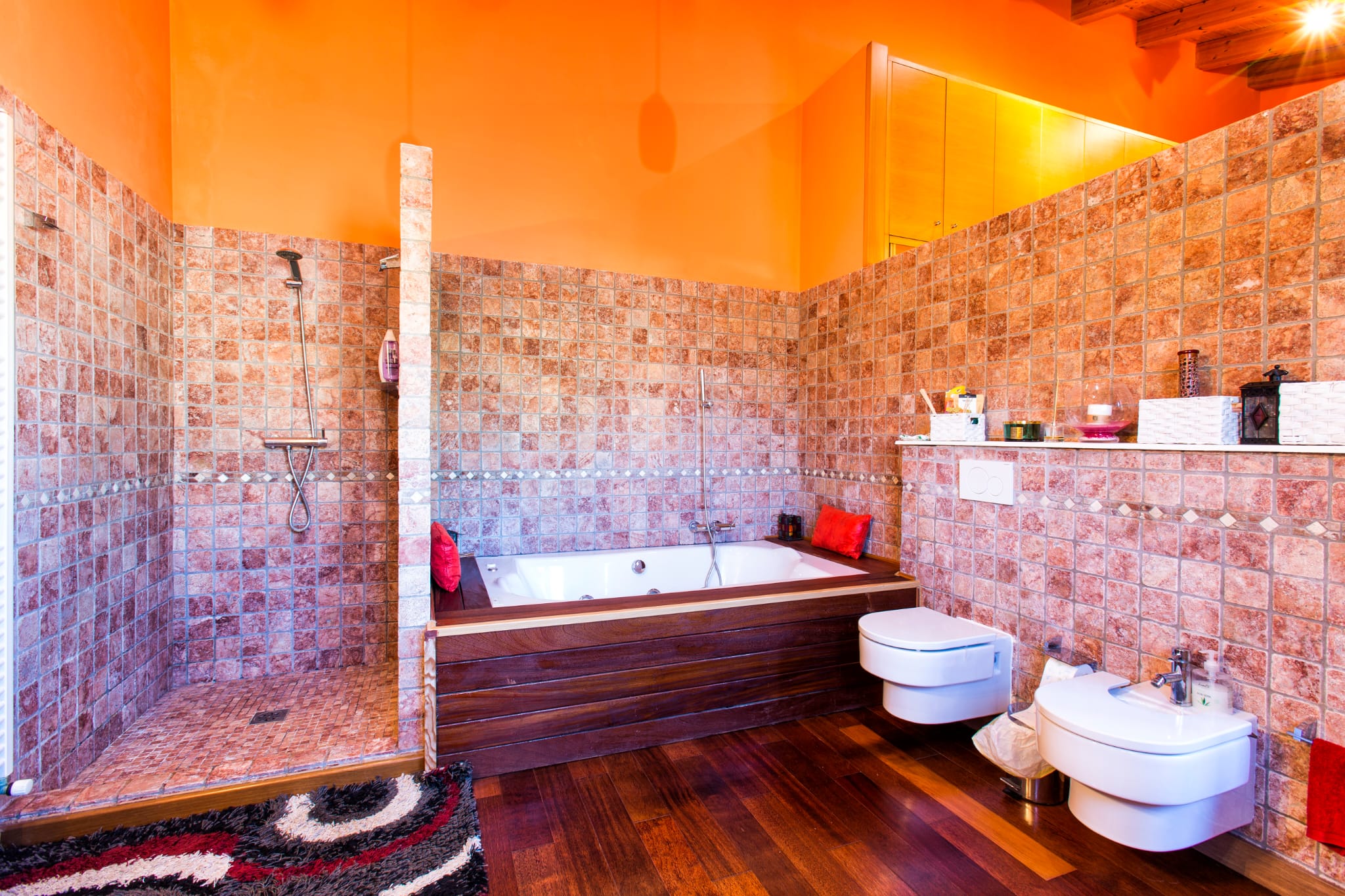 Badkamer met inloopdouche van natuursteen, een ligbad met donkerhouten ombouw en hangend sanitair. Oranje wanden contrasteren met de warme houten vloer.