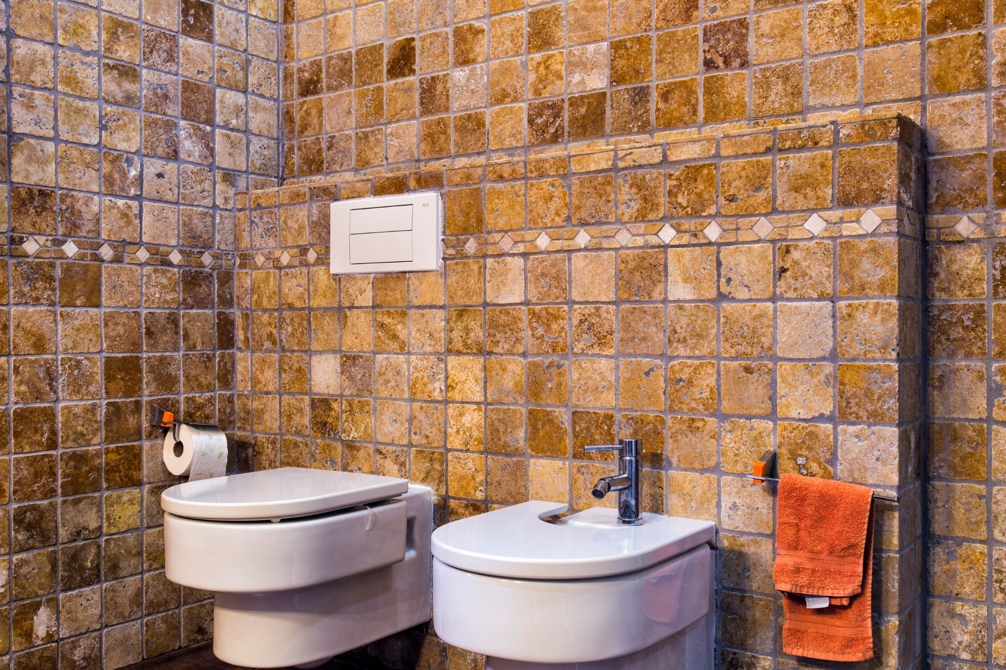 Badkamer met rustieke tegels in aardetinten, wandcloset en bidet van wit keramiek en een decoratieve mozaïekstrip.