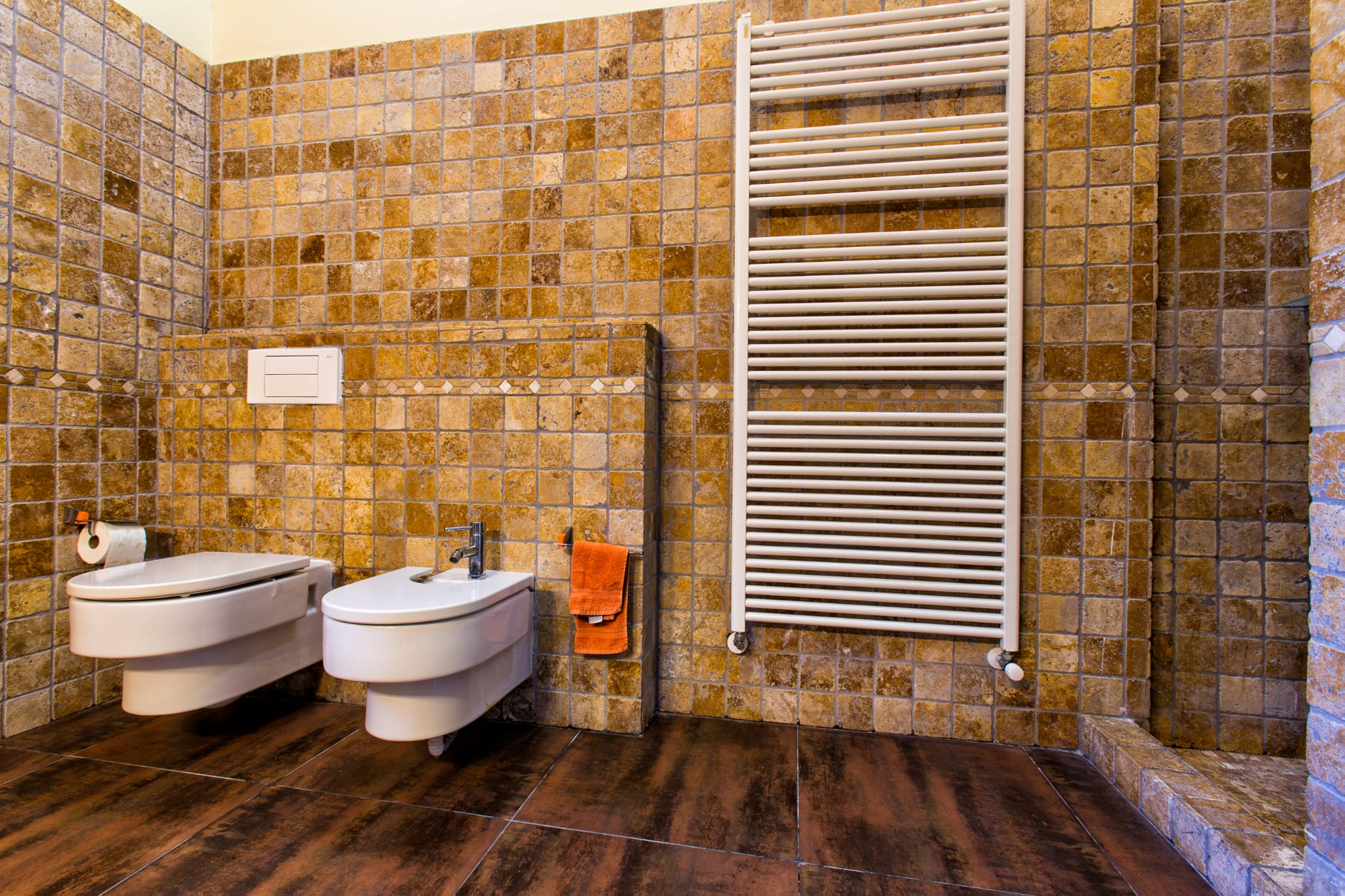 Badkamer met wandcloset en bidet, afgewerkt met aardetinten mozaïektegels en een grote witte handdoekradiator op een donkere tegelvloer.