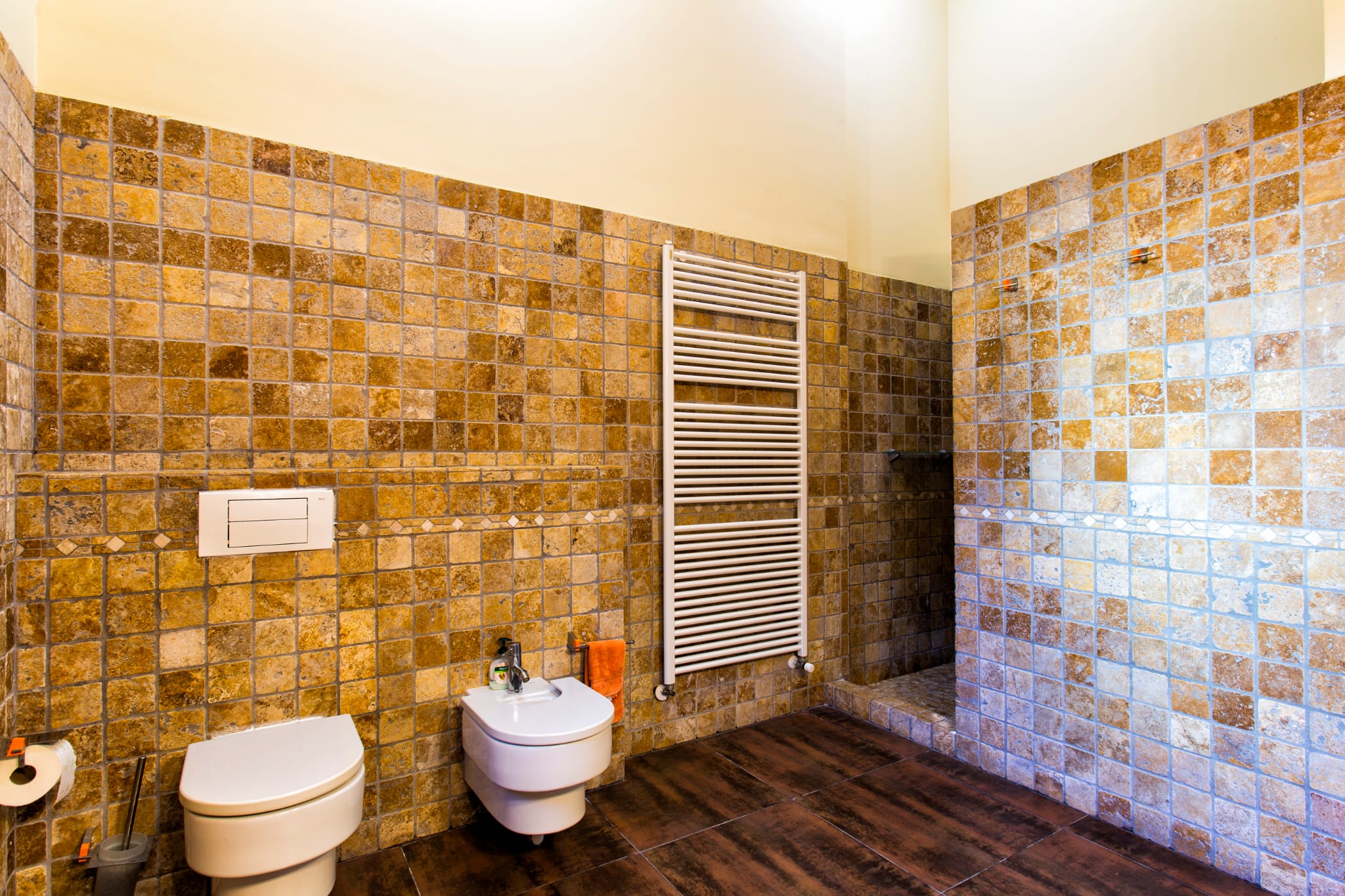 Badkamer met wanden van natuursteenmozaïek in aardetinten, hangend toilet en bidet, een witte handdoekradiator en een donkere houtlook vloer.