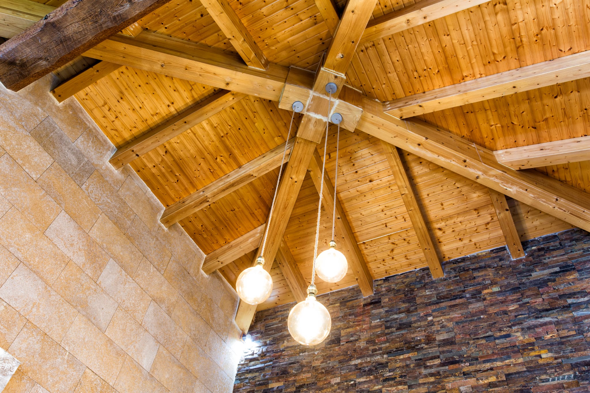 Detail van een houten balkenplafond in stervorm met drie hanglampen en muren van natuursteen en donkere leisteenstrips.