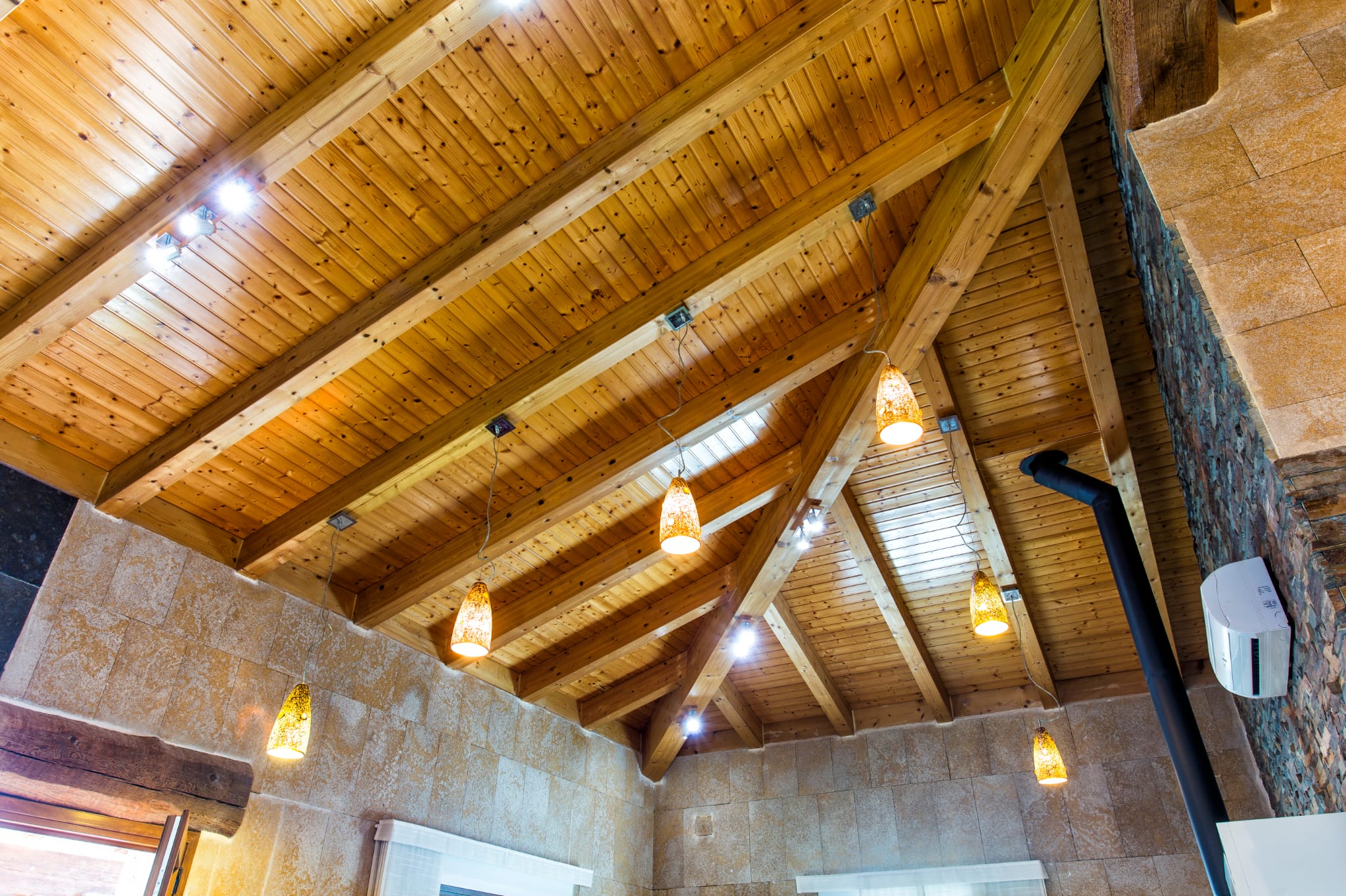Hoog gewelfd plafond met zichtbare houten balken en grenen panelen, verlicht door amberkleurige hanglampen en wanden van natuursteen.