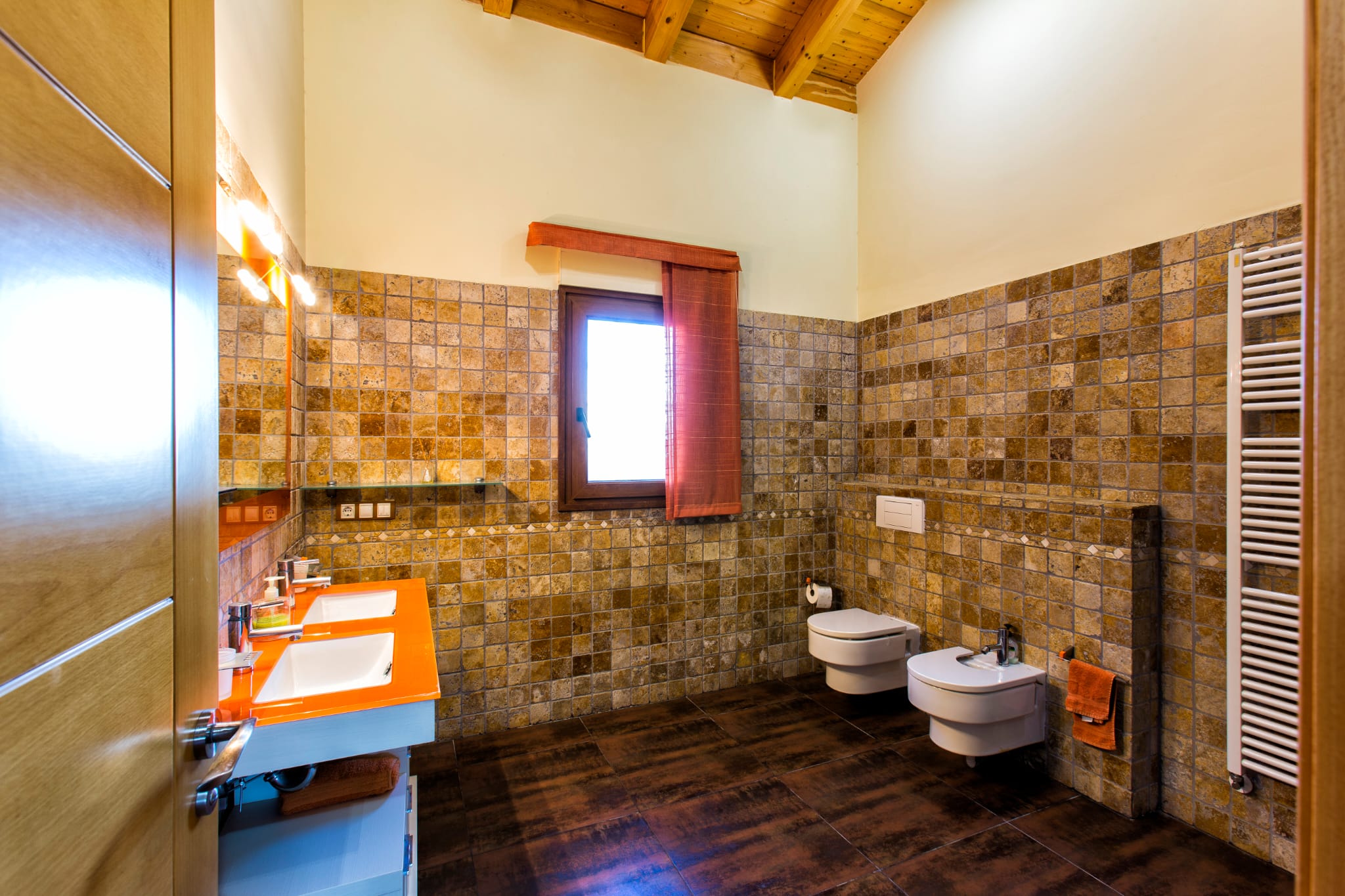 Moderne badkamer met dubbele wastafel met oranje blad, natuurstenen wanden en een gewelfd houten plafond met zichtbare balken.