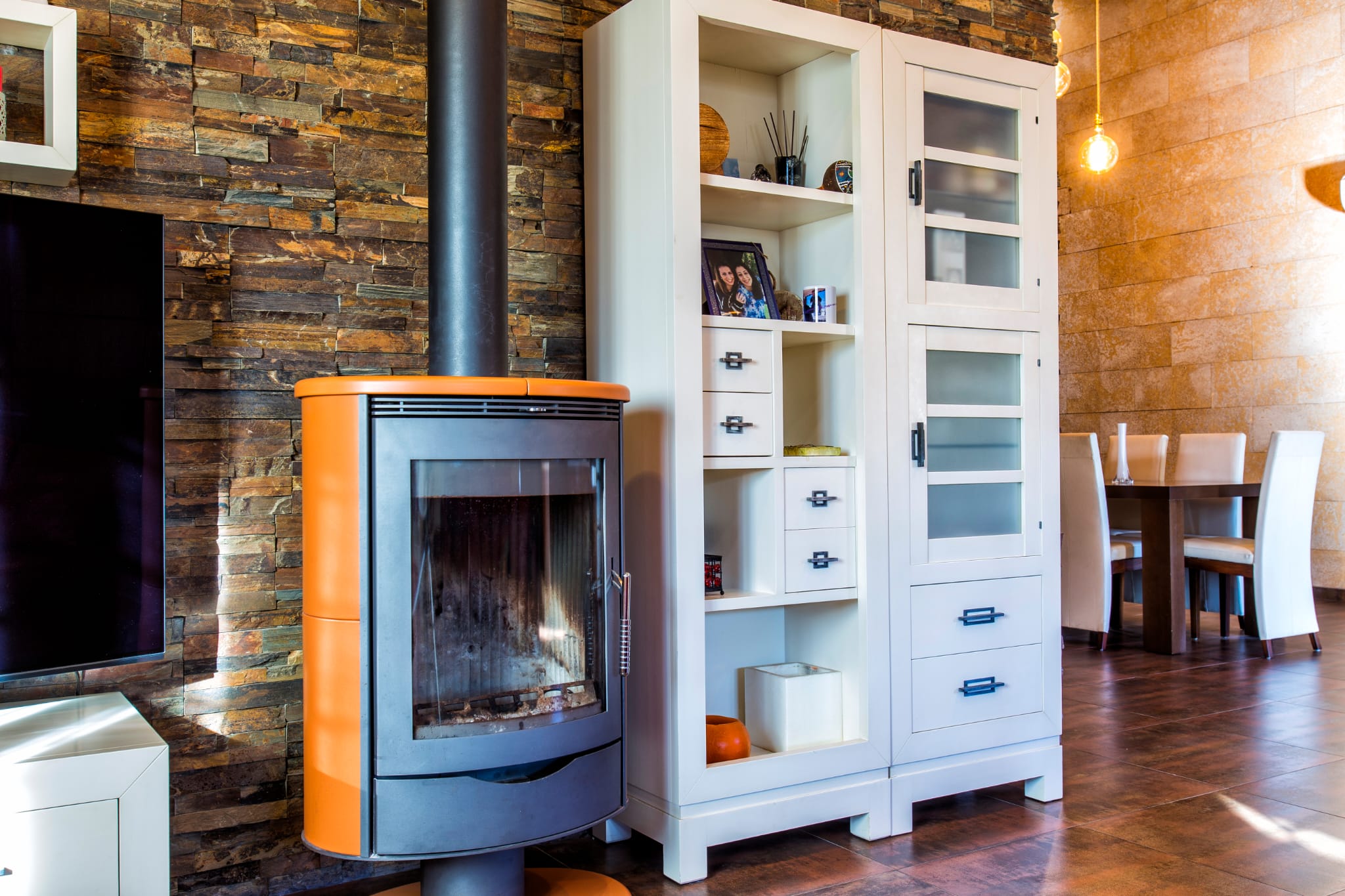 Moderne houtkachel met oranje details tegen een natuurstenen wand, geflankeerd door een witte kast en een eetgedeelte met tegelvloer.