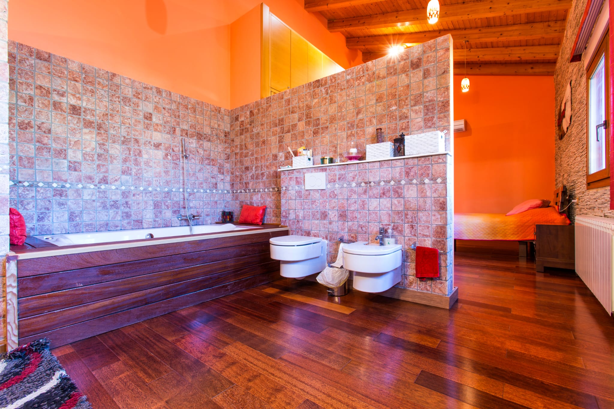 Open badkamer met een met hout bekleed bad, mozaïektegels en hangend sanitair, naadloos verbonden met de slaapkamer onder een rustiek houten balkenplafond.