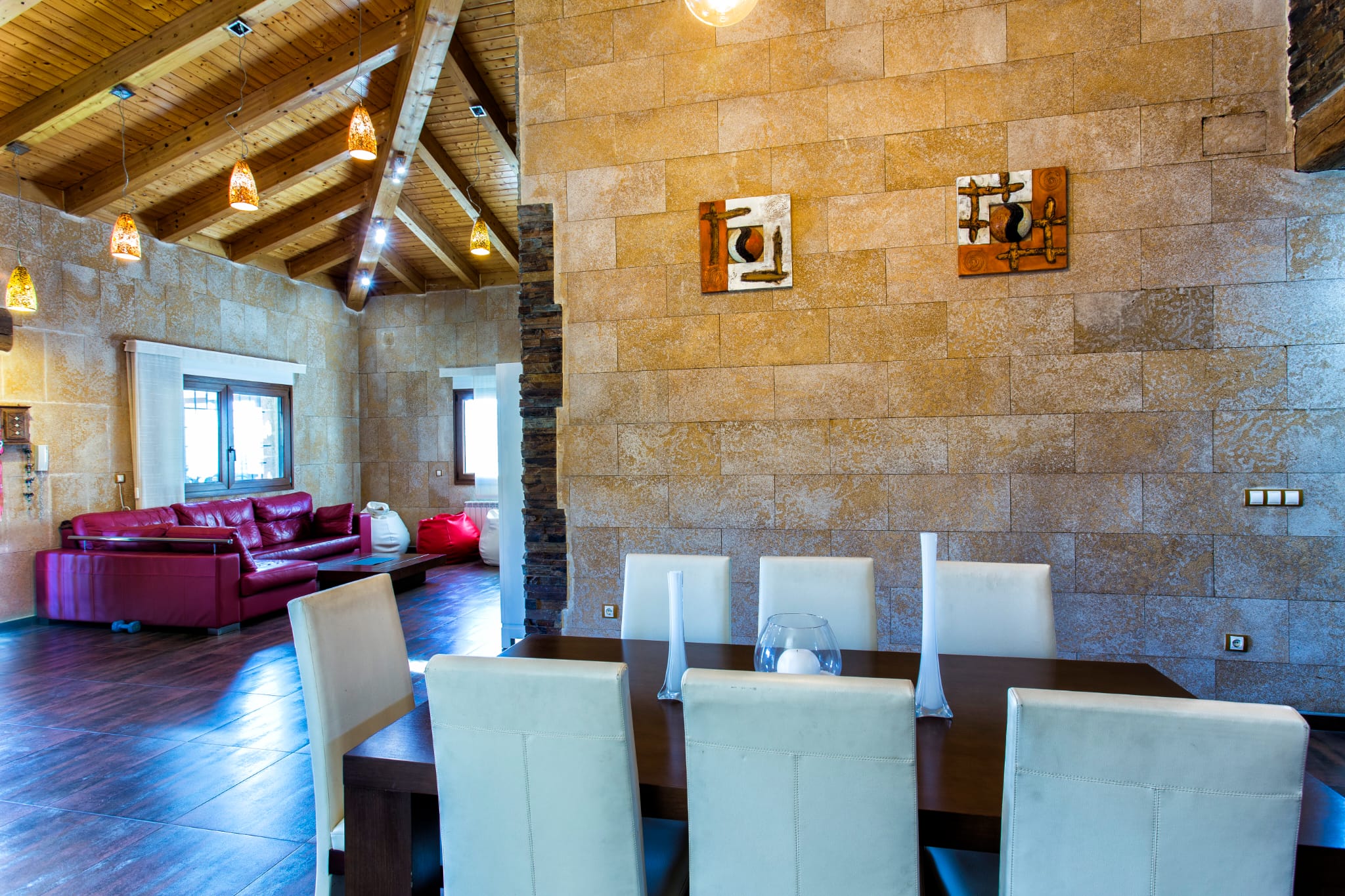Open eetkamer met natuurstenen wanden, een hoog houten plafond met zichtbare balken en een moderne eettafel die overloopt in de woonruimte met rode bank.