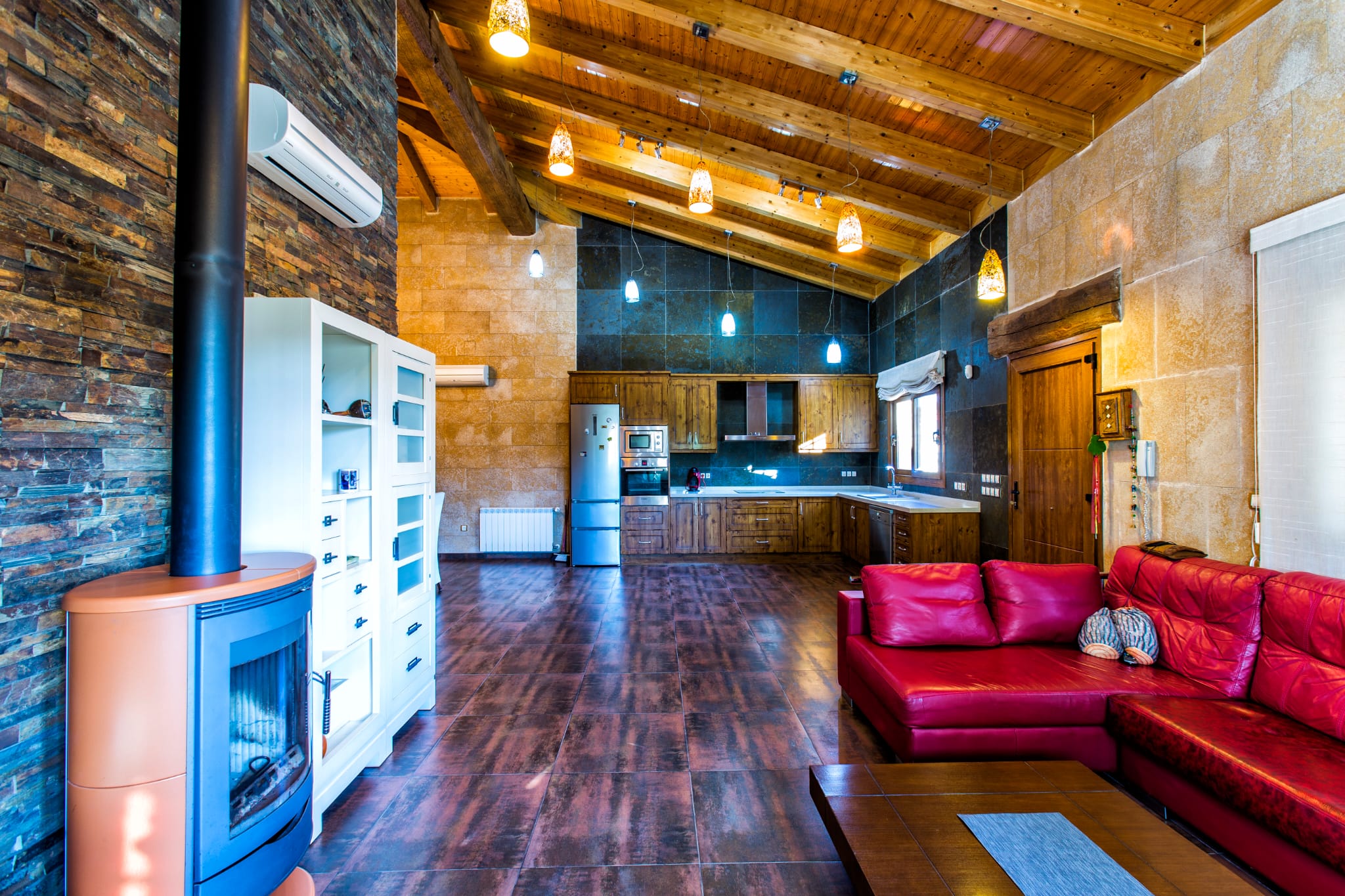 Open woonkamer met hoog houten balkenplafond, een moderne houtkachel en rustieke keuken met inbouwapparatuur, afgewerkt met natuurstenen wanden en plavuizen.