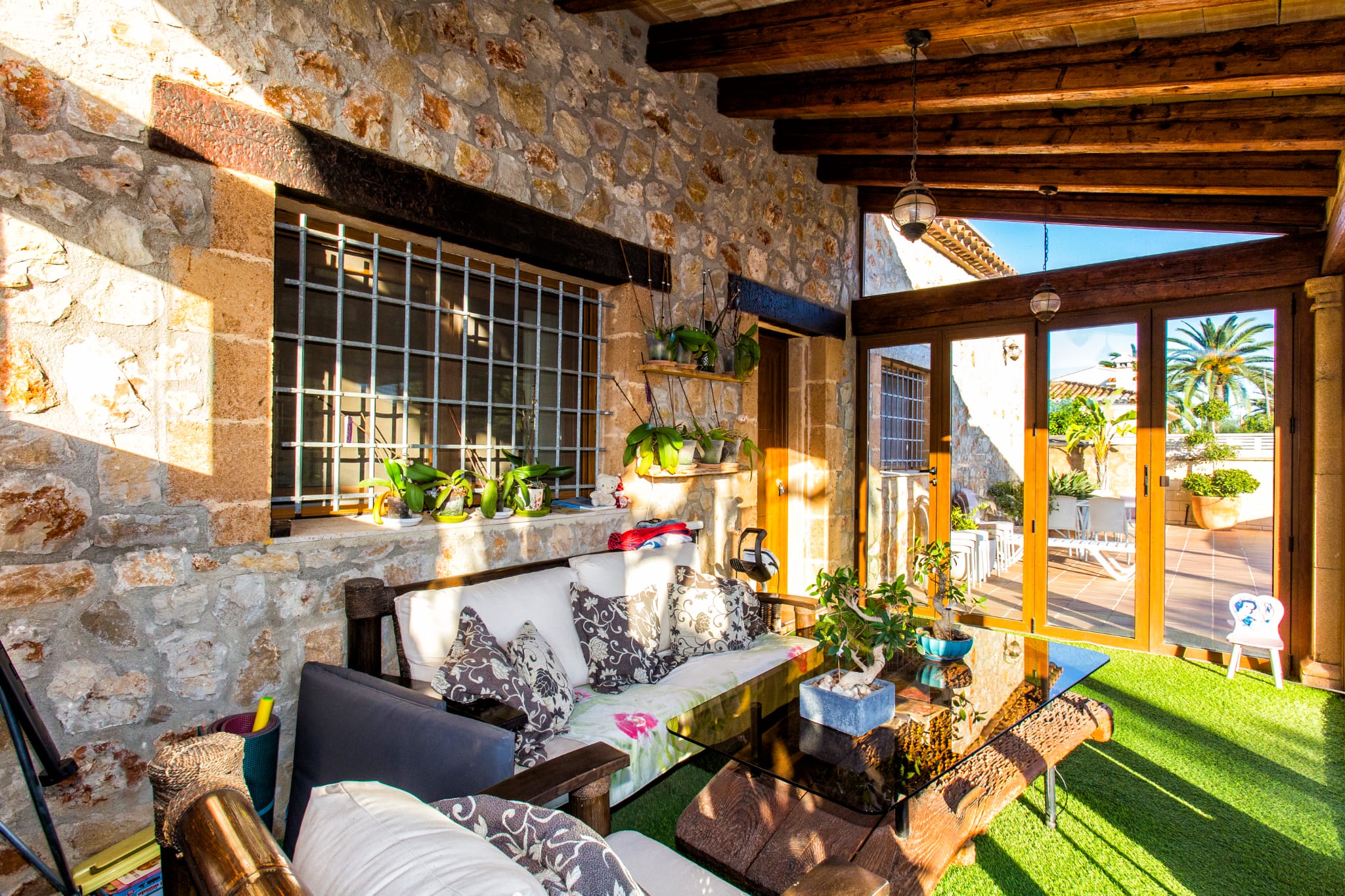Overdekt terras met natuurstenen muren, donkerhouten balken en zithoek met glazen tafel op kunstgras, uitkomend op een zonnige patio.