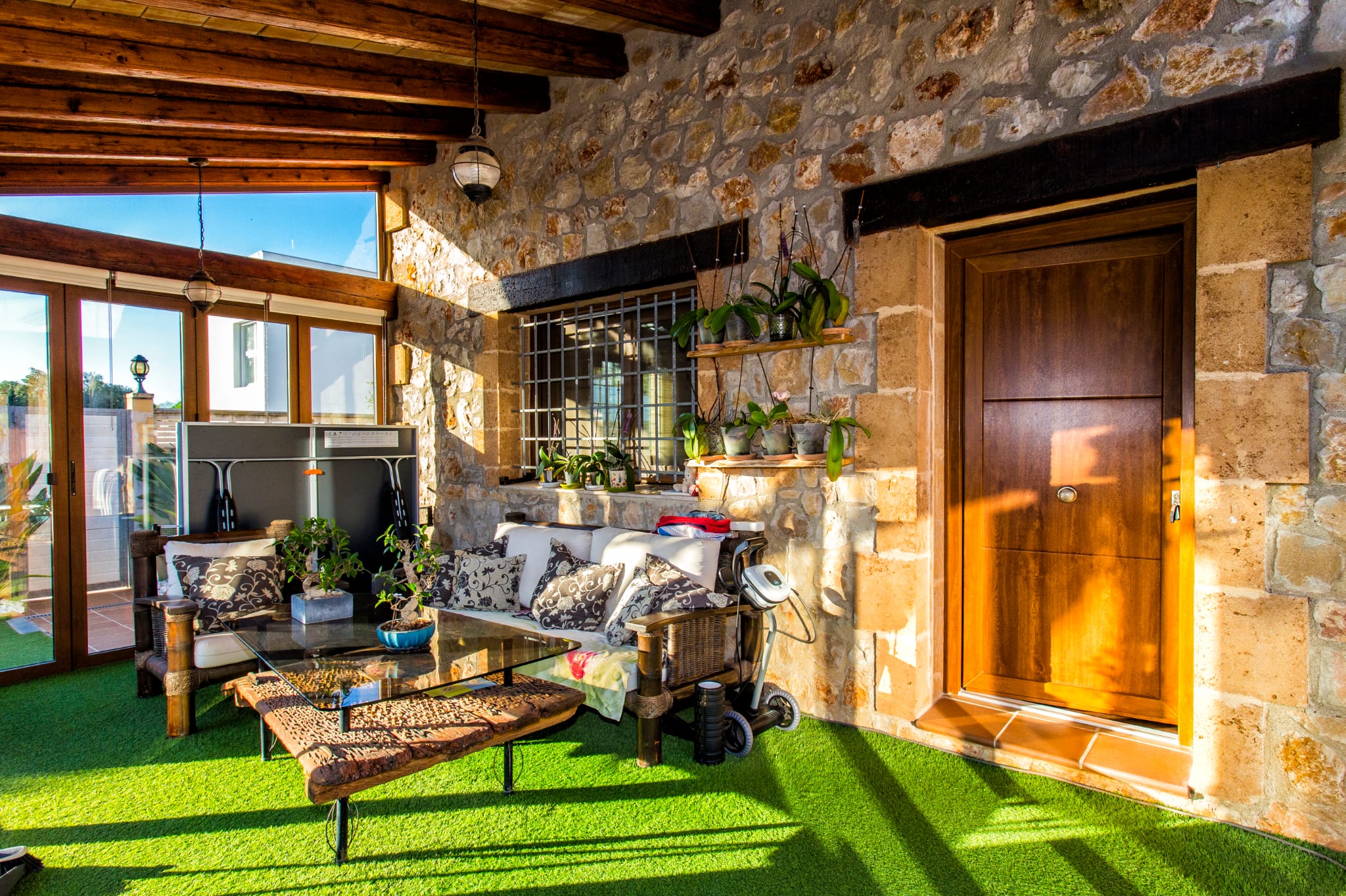 Overdekt terras met natuurstenen muren, houten plafondbalken en kunstgras. Voorzien van een zithoek met een salontafel van drijfhout en orchideeën.
