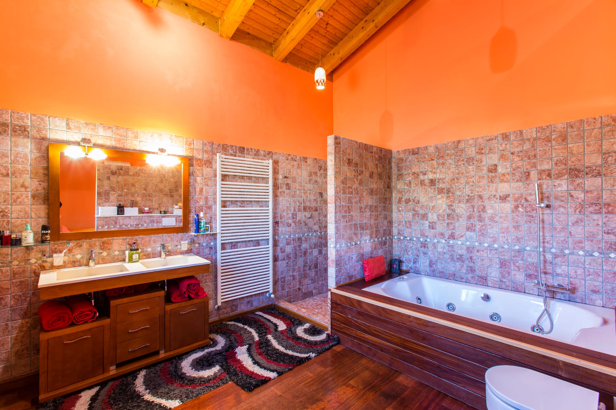 Rustieke badkamer met houten balkenplafond, dubbele wastafel op een donkerhouten meubel, whirlpool bad en wanden in warme terracotta- en oranjetinten.