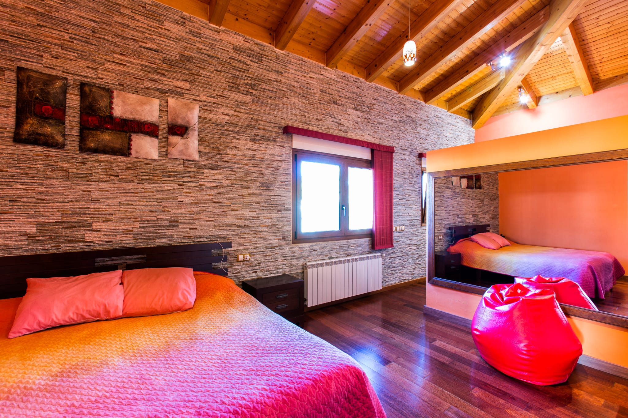 Slaapkamer met een getextureerde stenen wand, gewelfd houten balkenplafond en donkere vloeren. Een grote wandspiegel reflecteert het bed met roze en oranje textiel.