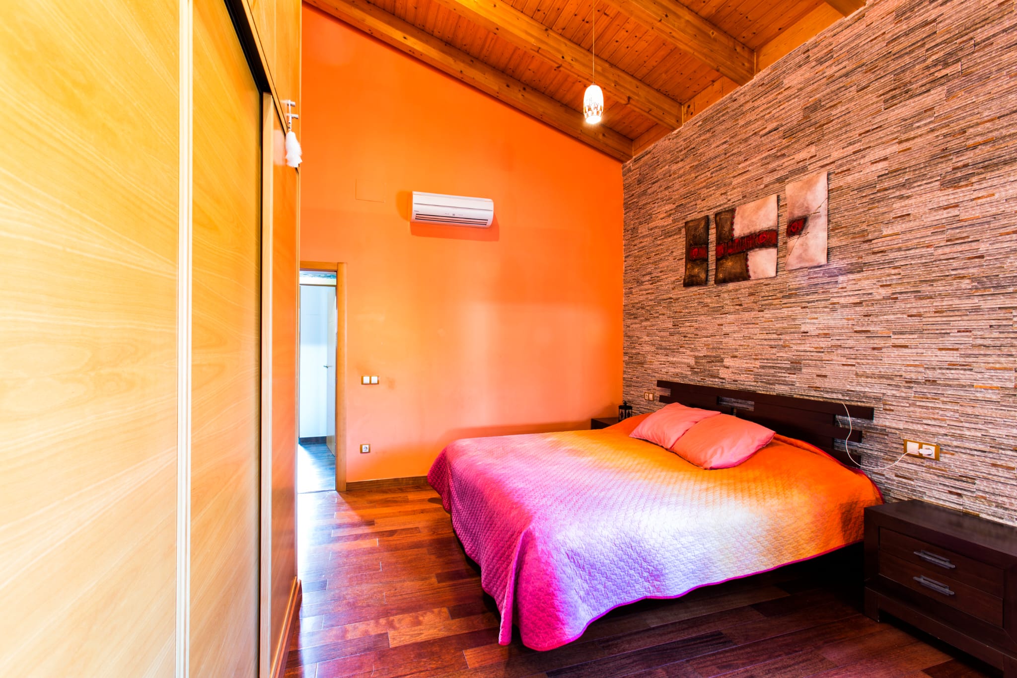Slaapkamer met een schuin houten balkenplafond, een natuurstenen accentmuur en inbouwkasten. Voorzien van een houten vloer, airconditioning en donkerhouten meubilair.