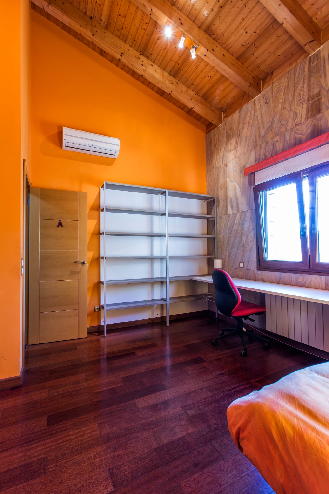 Slaapkamer met hoog houten balkenplafond, oranje muren, een geïntegreerde werkplek met bureau, metalen stellingen en een donkere houten vloer.
