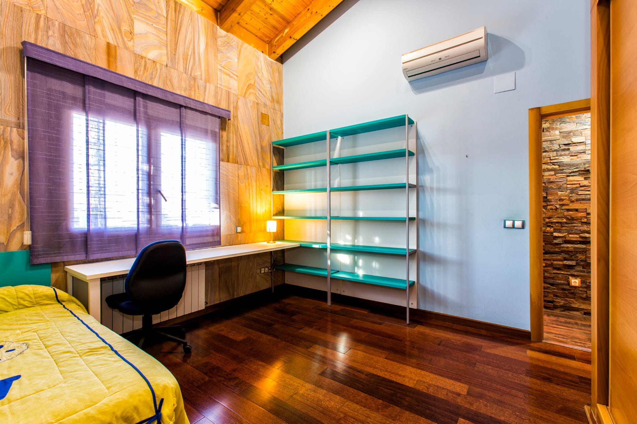Slaapkamer met werkruimte, schuin houten plafond, turquoise stellingkast en wit bureau onder een raam met paarse jaloezieën.
