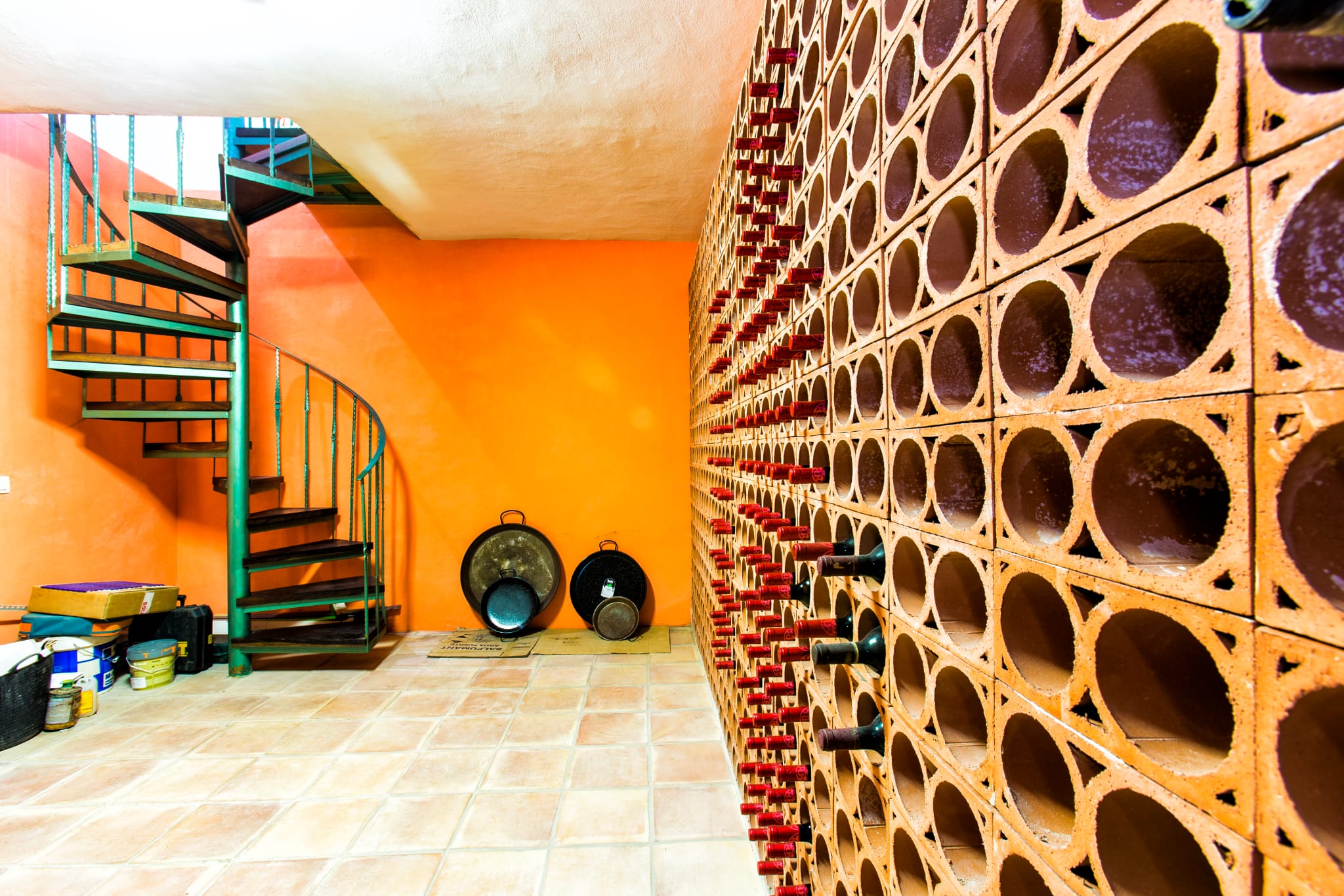 Wijnkelder met een terracotta flessenrek, oranje accentmuur en een groene metalen wenteltrap voorzien van donkere houten treden.