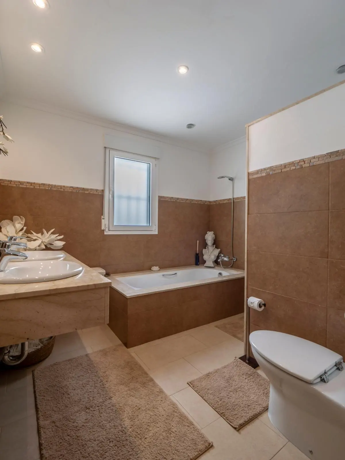 Badkamer met dubbele wastafel op marmeren blad, ingebouwd ligbad met bruine tegels en inbouwspots in het plafond.