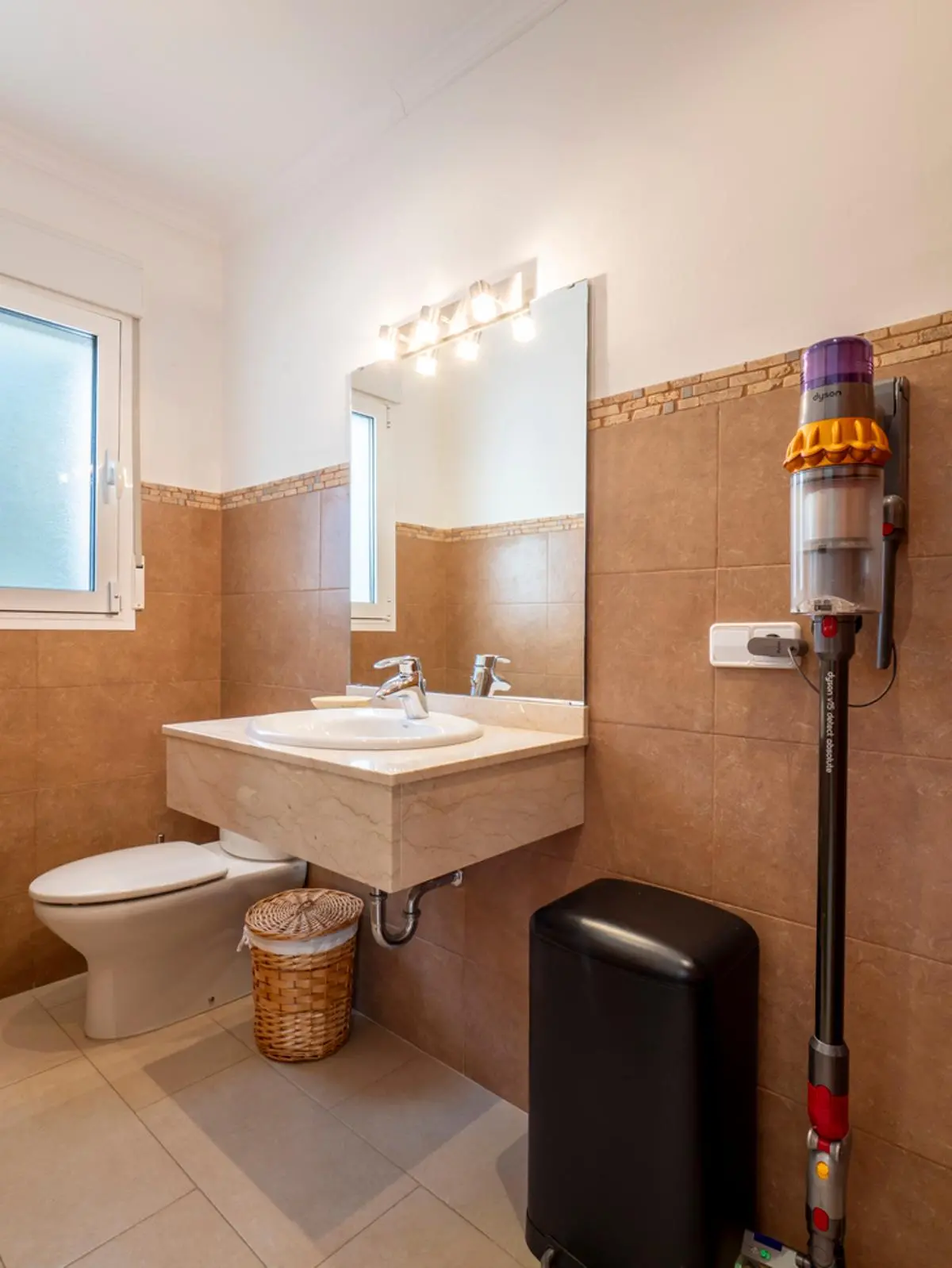 Badkamer met zwevend marmeren wastafelmeubel, beige wandtegels en een grote spiegel met verlichting. Voorzien van daglicht via een raam en praktische details.