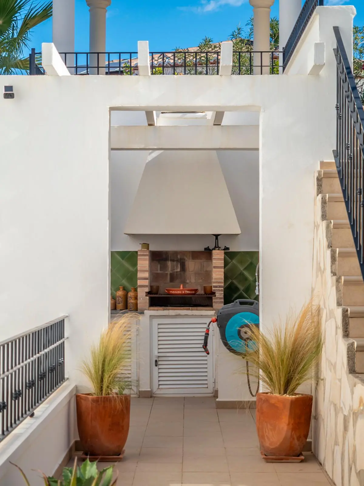 Buitenbarbecue met bakstenen grill, witte schouw en groene tegelwand, geflankeerd door terracotta plantenbakken en witte muren.
