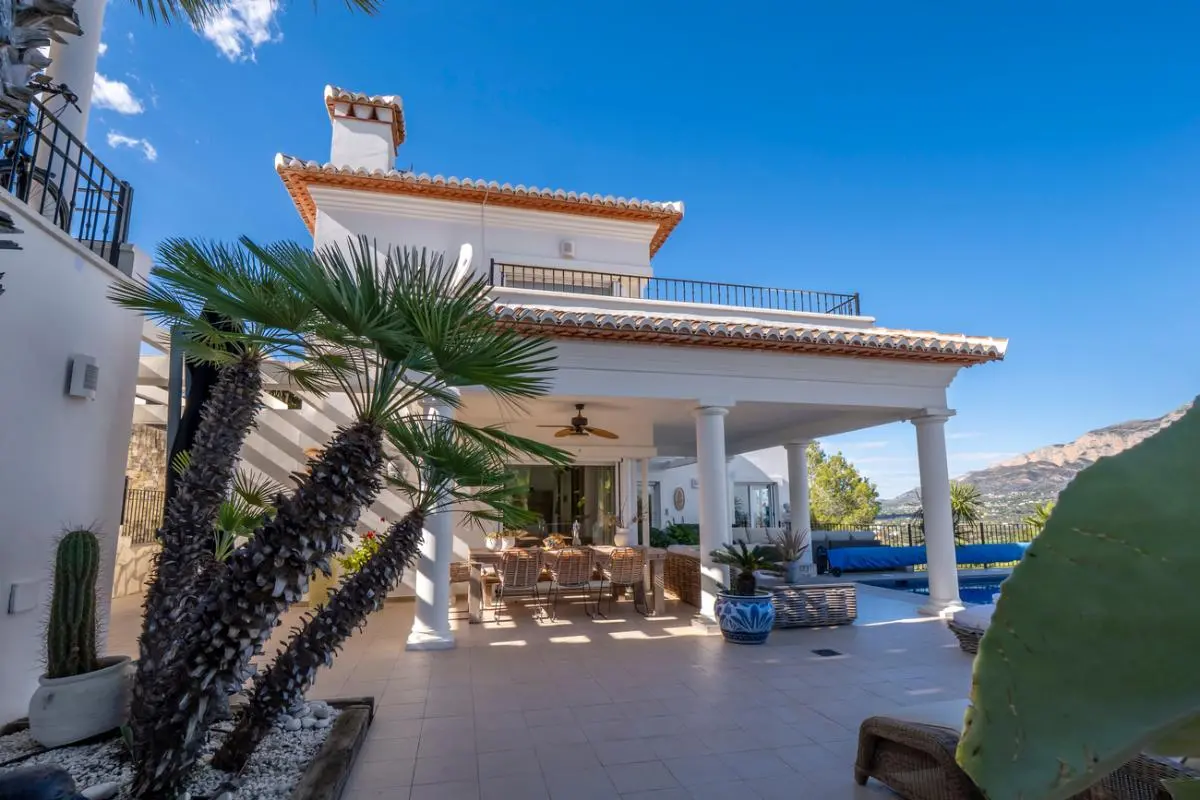 Exterieur van een witte villa met een overdekt terras met zuilen, een eethoek in de buitenlucht en een mediterrane tuin met uitzicht op de bergen.