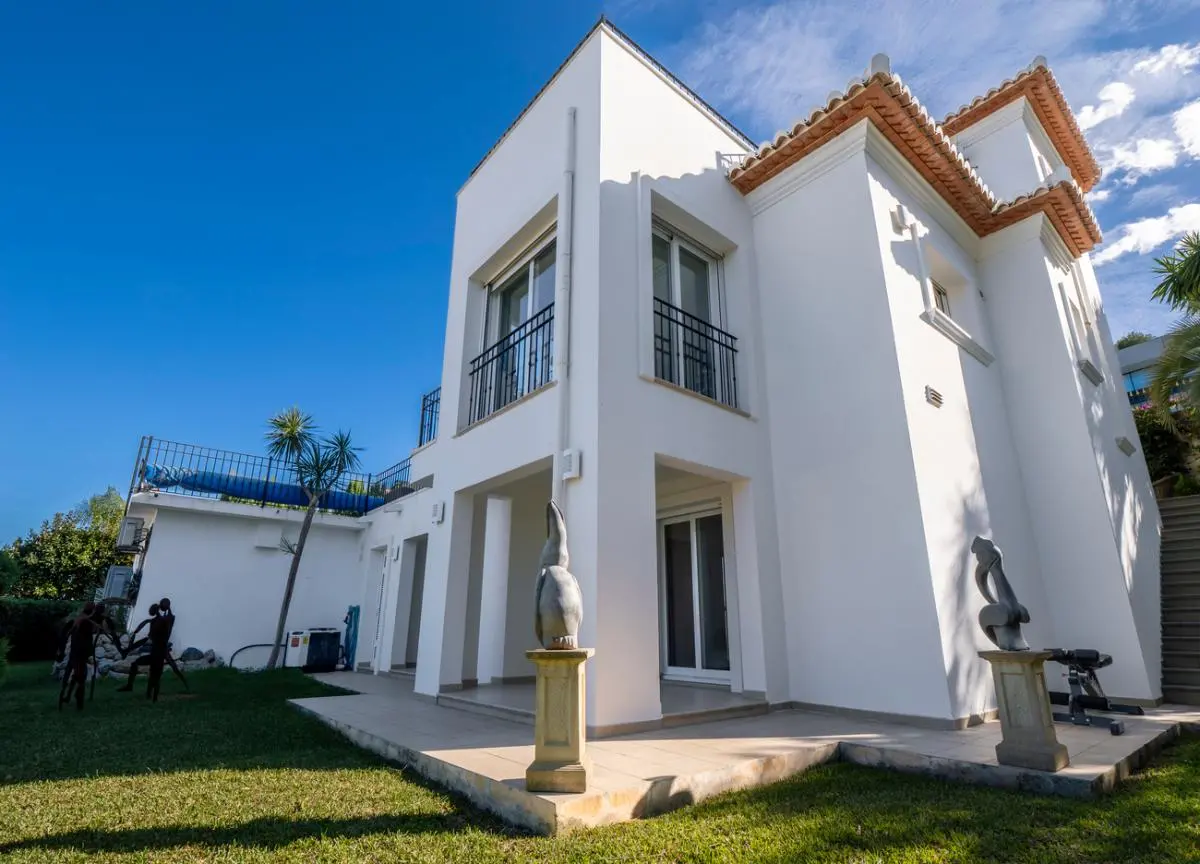 Exterieur van een witte villa met terracotta dakpannen, zwarte ijzeren balustrades en een gazon voorzien van decoratieve stenen sculpturen op sokkels.