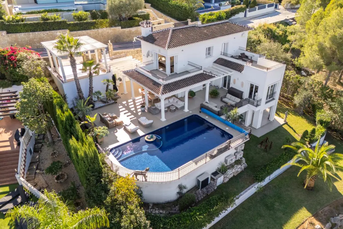 Luchtfoto van een witte mediterrane villa met een groot zwembad, meerdere overdekte terrassen met zuilen en aangelegde tuinen met palmbomen.
